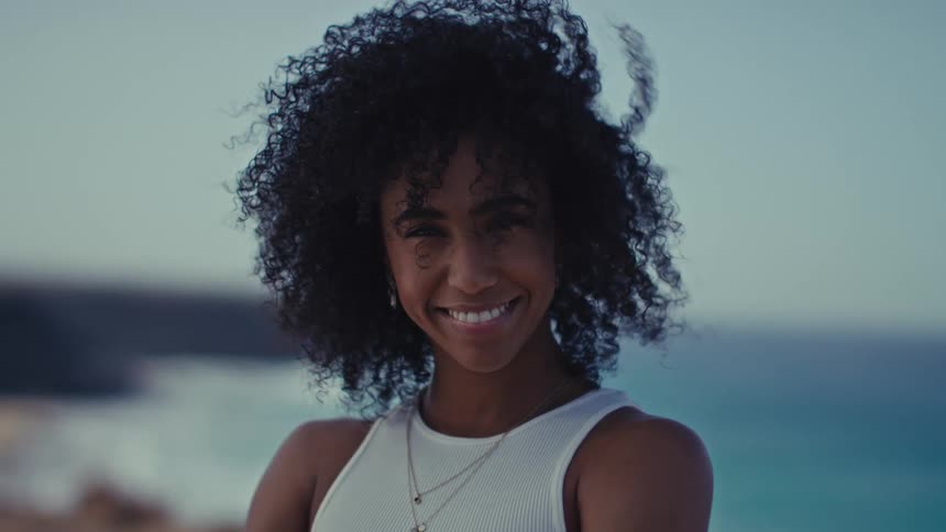 A woman with dark curly hair smiles while standing outdoors near the ocean at dusk the wind gently blows through her hair as she looks directly into the camera