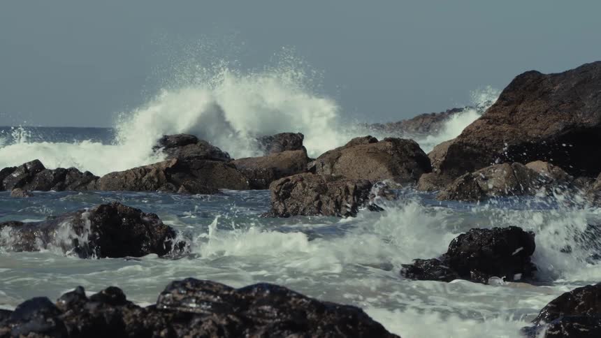 Heavy waves crash against the rocky shoreline at midday breaking into bright scattered splashes as they strike the sunlit rocks