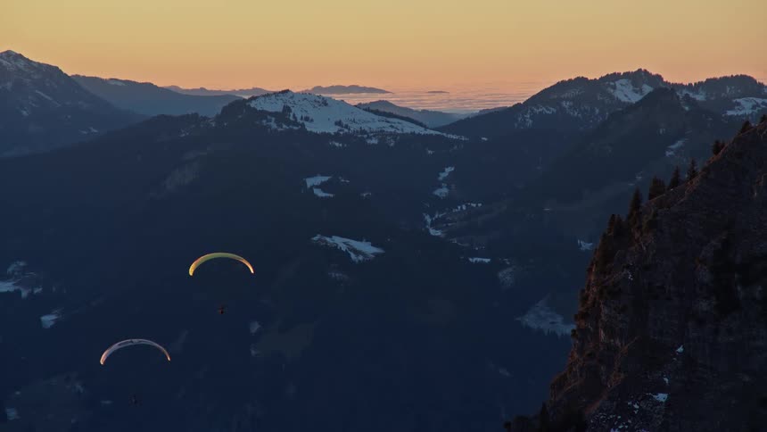 Two paragliders with glowing canopies drift through a dark mountain valley at dusk with snowcapped peaks and a soft orange glow on the horizon in the background