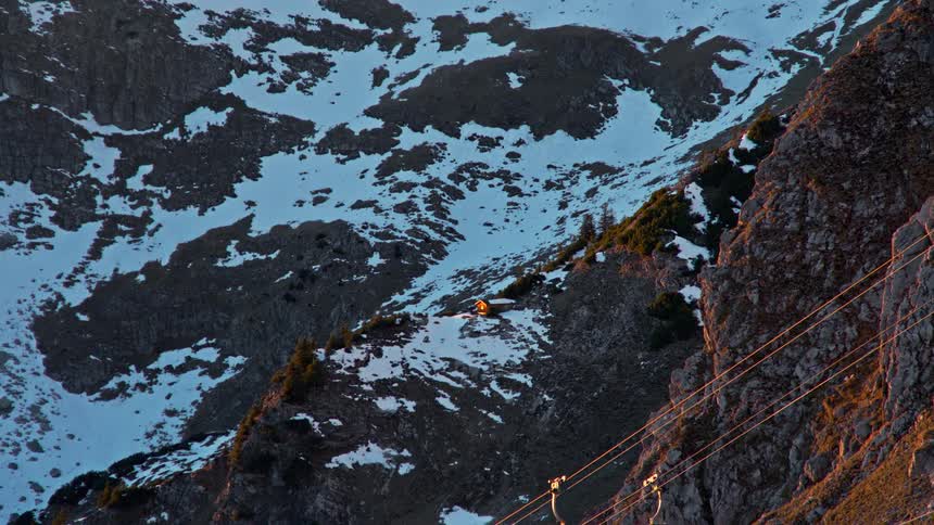 A cable car glides down a steep rocky mountain slope partially covered in patches of snow warm sunlight illuminates the side of the cliff and the cable car as it descends into the shadowed valley below