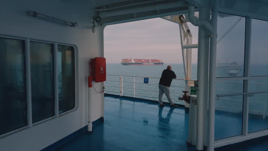 A man leans against a ship railing while watching a cargo ship in the distance