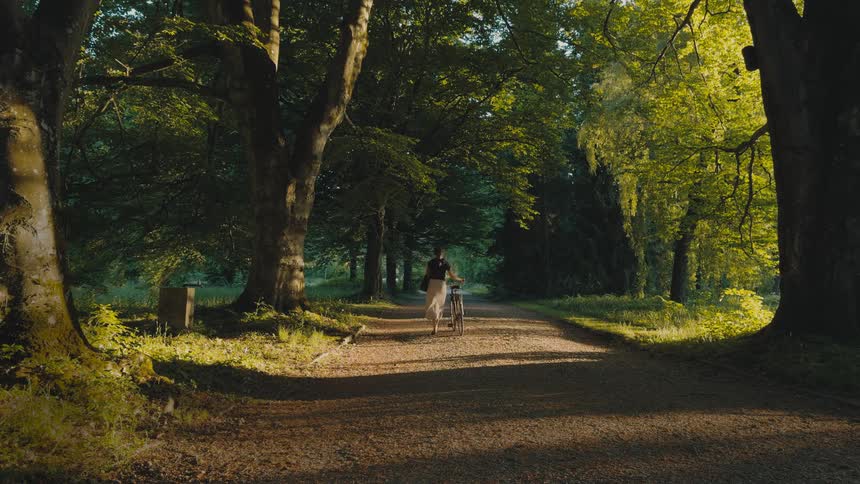 A woman walks her bicycle along a sunlit forest path surrounded by lush greenery