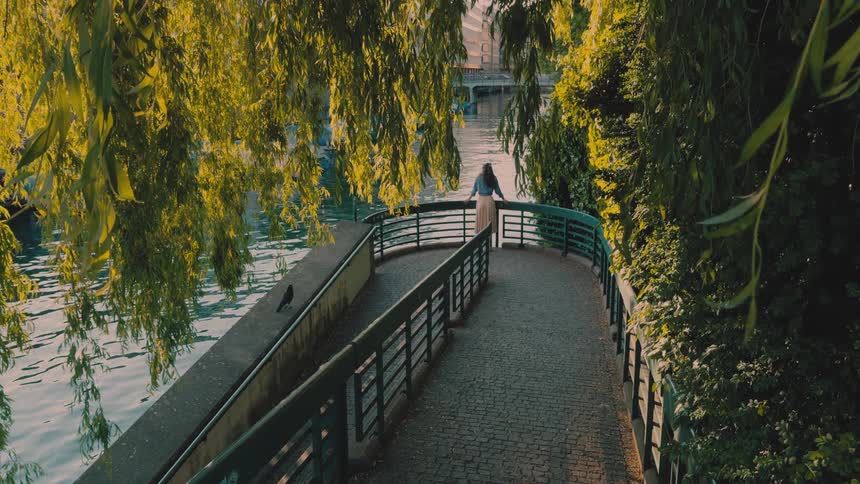 A woman stands at the end of a curved riverside walkway framed by hanging green foliage as sunlight reflects off the water