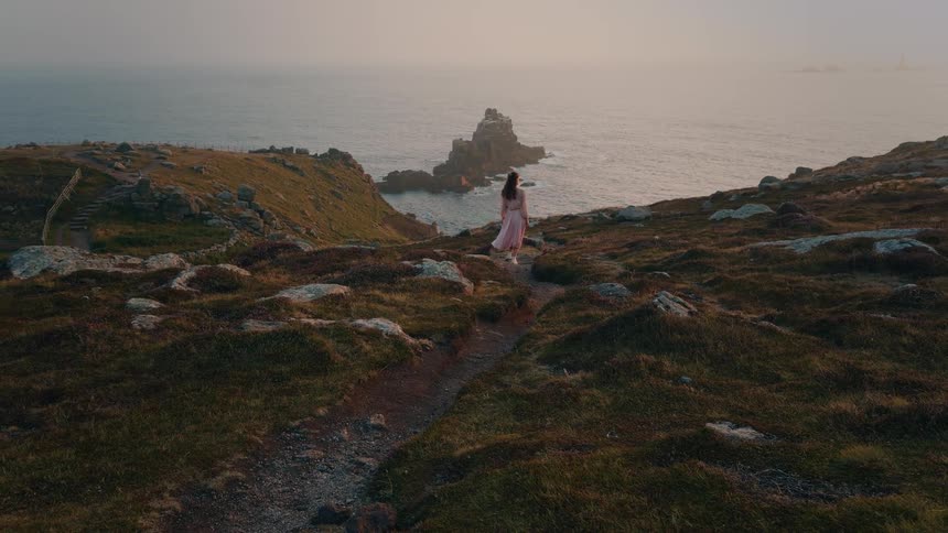 Woman wearing a long skirt walks on a mountain path to the coast