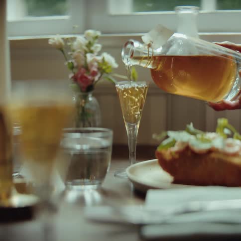 At a beautifully set dining table near a window a hand pours a golden tea from an elegant decanter into a delicate tall glass beside a meal