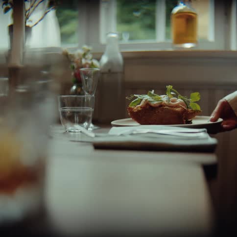 A persons hand places a white plate holding a thick slice of toast topped with lightcolored meat and fresh green herbs onto a wooden table set with a glass of water and a napkin