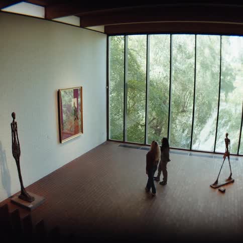 Two figures walk across a polished wood floor in a sunlit gallery passing thin elongated sculptures as they head toward a massive glass wall overlooking a lush green foresta large framed painting hangs on a white brick wall contrasting with the natu