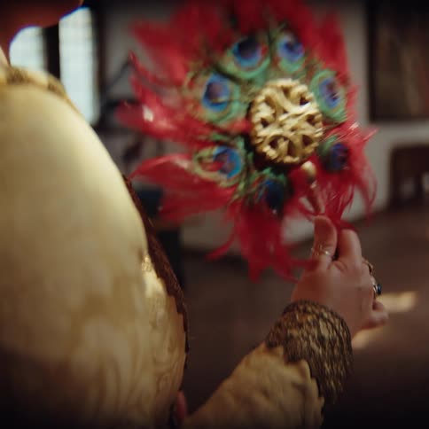 A person in periodstyle clothing holds an ornate fan decorated with red feathers and peacock eyesthe fan features a gold filigree center and is being gently twirled between the persons fingers
