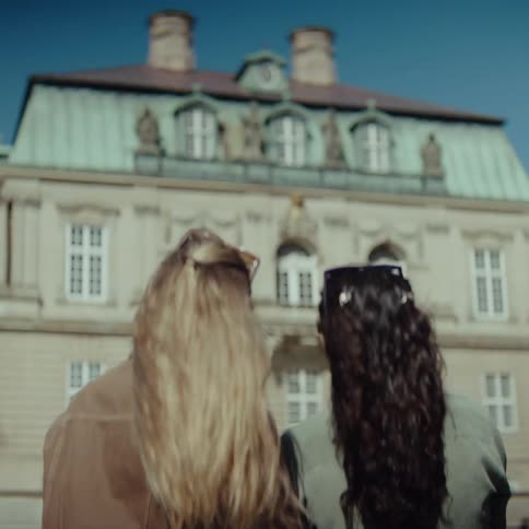 Two women one with blonde hair and one with dark curly hair stand with their backs to the camera as they look toward a large ornate historic building with a light green roof and many windows