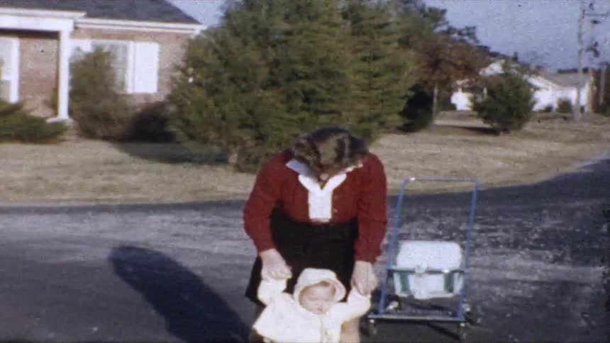 Young adult woman holding the hands of a baby trying to walk in front of a house in a suburban area outdoors during day time