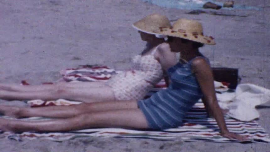 Two women wearing straw hats and bathing suits are sitting on a striped towel on a sandy beach