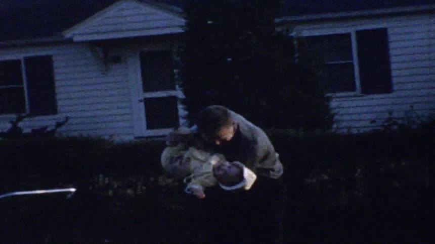 A man gently swings a baby dressed in white down toward the ground in the dark yard outside a house