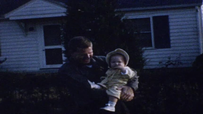 A man stands in a yard at dusk holding a small baby dressed in a yellow outfit and bonnet and they are positioned in front of a white house with dark shutters and a large evergreen tree