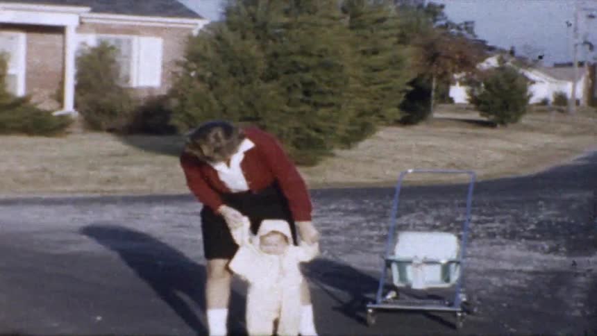 Person helping a baby walk on a residential street with a stroller nearby