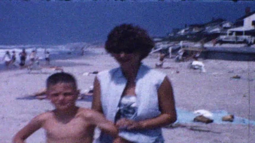 A woman with short dark hair and a young boy stand on a crowded sandy beach in front of coastal houses while people lounge on towels and the ocean waves crest in the background under a clear blue sky during a sunny day