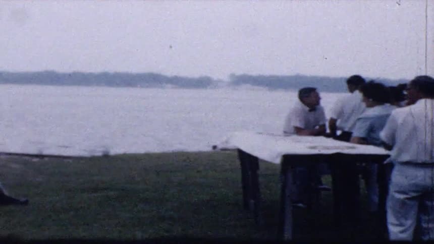 Some people gathers by a lake on a grassy lawn where a man relaxes in a lounge chair near a toddler and dog while other people socialize around a picnic table under the trees