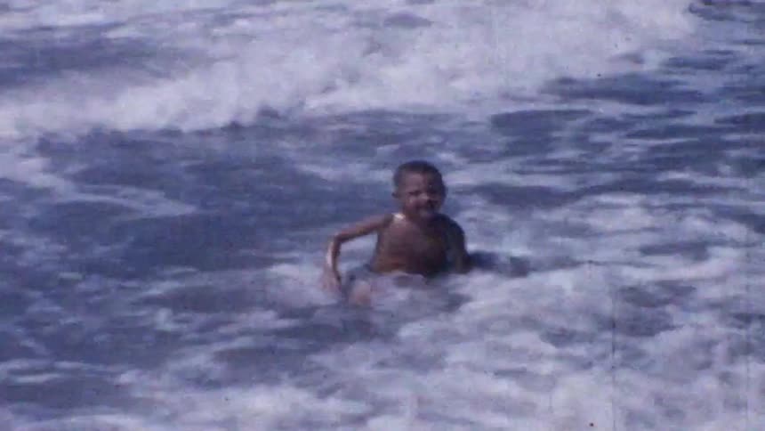 A young boy in swim trunks enjoys the ocean waves as they wash over him on a sunny beach while other people wade in the background
