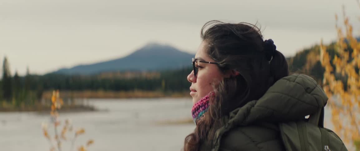 Woman wearing glasses and a jacket looking over a river with forest and mountains in the background