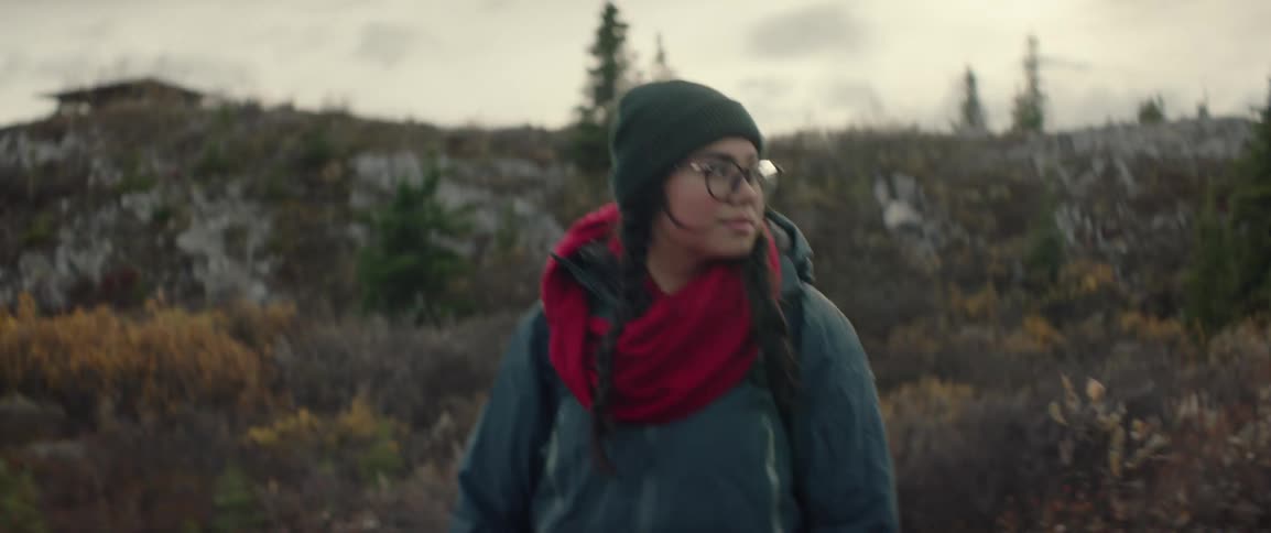 A woman wearing a dark beanie glasses and a vibrant red scarf walks through a rugged autumnal outdoor landscape