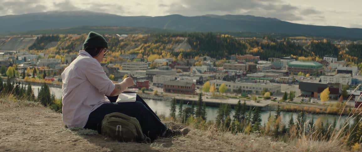 A woman sits on a dirt ridge overlooking a remote town and mountains while painting