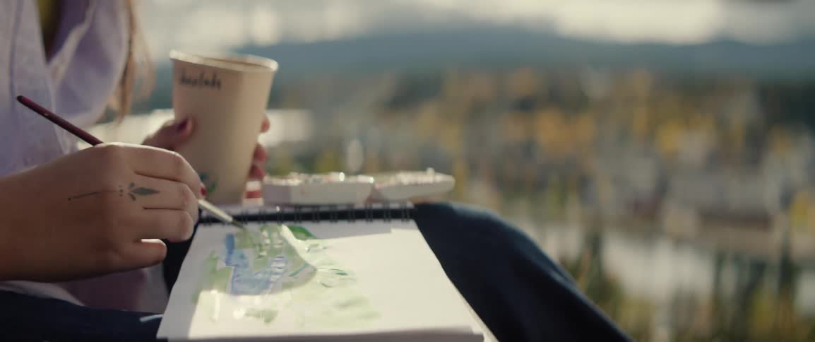 Person paints a landscape in a sketchbook while holding a coffee cup outdoors
