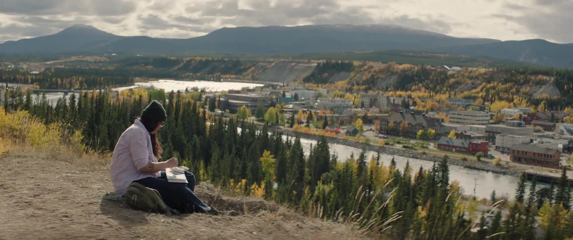 A woman paints in a watercolor sketchbook while sitting on a hillside overlooking a town in the afternoon sun