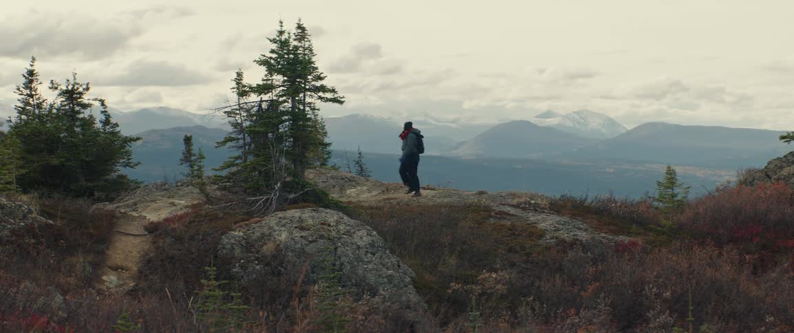 A person wearing a backpack and a red scarf walks across a rocky highaltitude ridge toward some evergreen treesthe background reveals a vast mountain range under a cloudy sky overlooking a valley with distant peaks