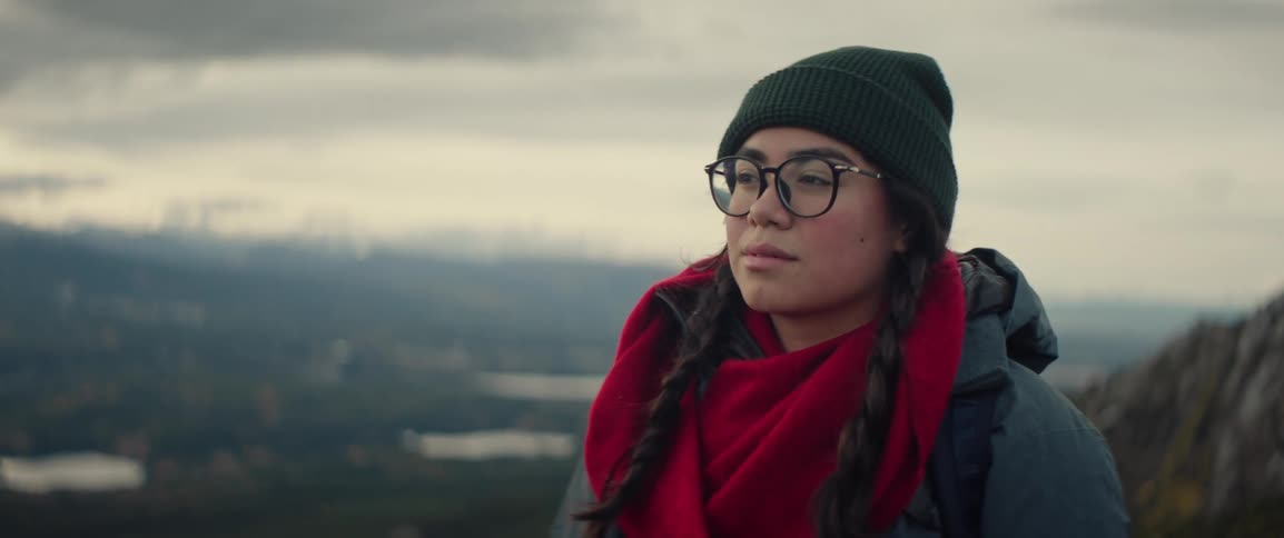 Woman wearing a beanie glasses and a scarf overlooking a valley landscape under a soft sky
