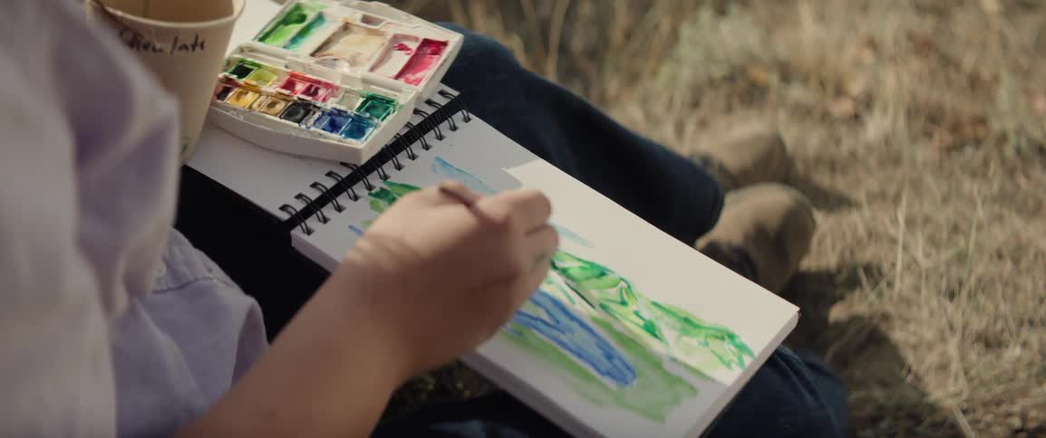 An artist paints a watercolor landscape of green mountains and a lake while outdoors