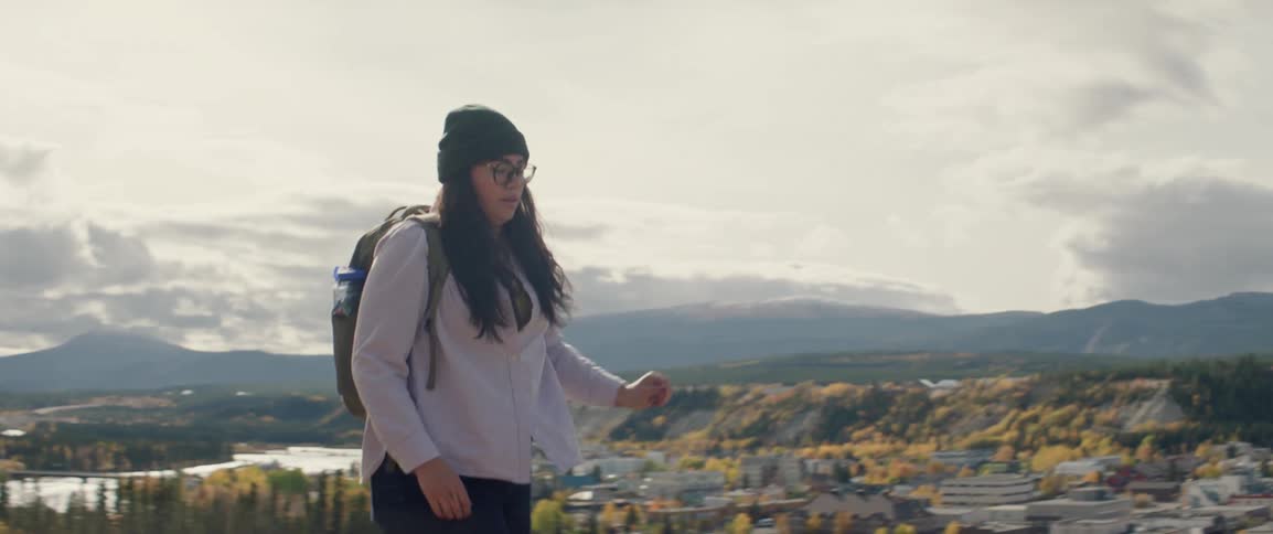 Woman walking along an elevated trail overlooking a forested valley river and downtown Whitehorse in the background