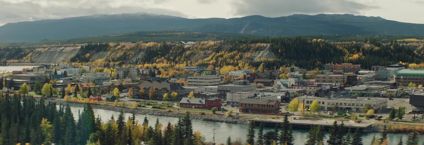 A highangle shot shows a town nestled between a river and rolling hills covered in yellow autumn trees under an overcast sky