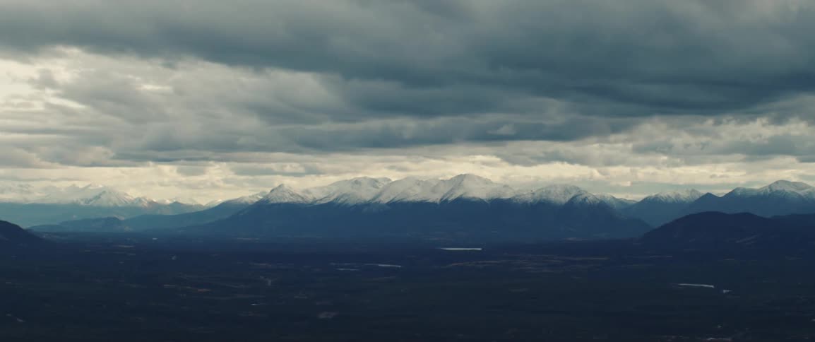 Mountain range with snowcapped peaks under heavy clouds and a vast plain in the foreground
