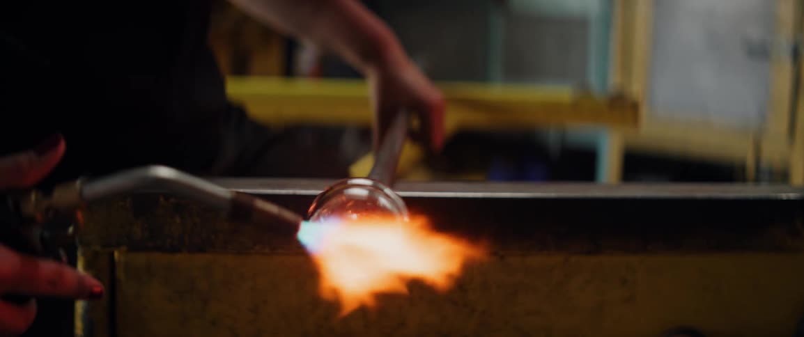 A worker uses a blowtorch to heat and shape molten glass as bright flames flare in an industrial workshop