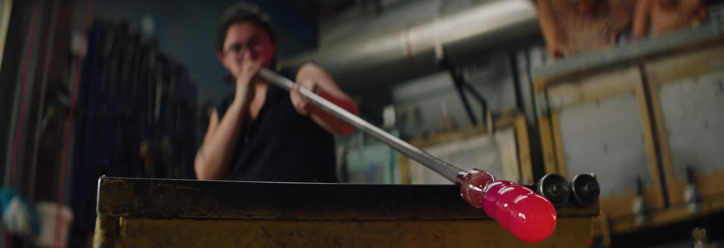 A young woman works inside a workshop blowing into a piece of molten glass to shape it