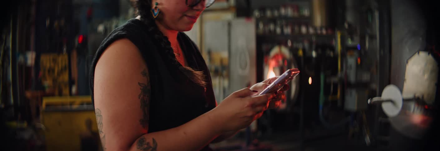 Person using a smartphone in a glassblowing workshop with tools in the background