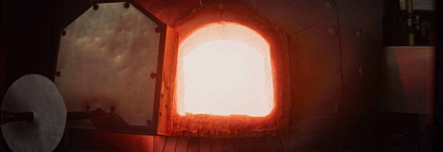 A persons hand pulls open the door of an industrial furnace inside a workshop to reveal intense heat