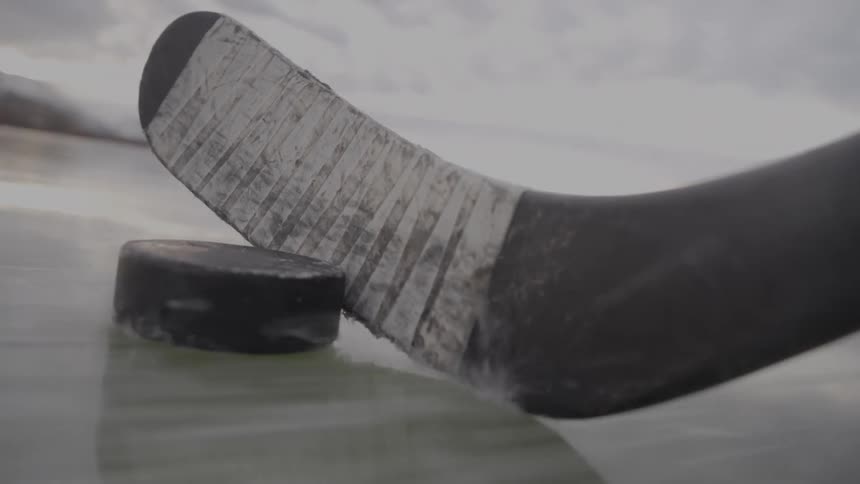 A hockey stick wrapped in white tape handles a black puck across a frozen lake showing the reflection of the equipment on the natural ice surface with a blurred mountain range and a cold overcast sky visible in the background