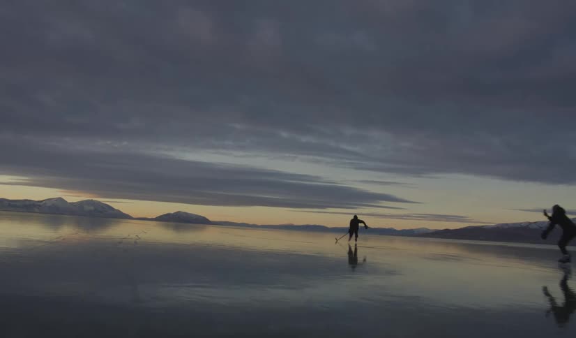 Two people play ice hockey on a wide frozen lake at dusk skating with sticks and a puck across the mirrorlike ice beneath a dramatic sky