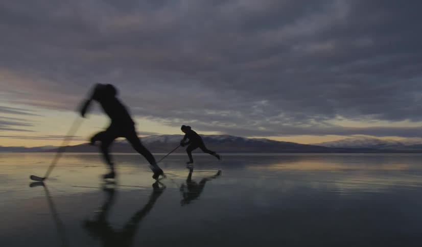 Two people skate on a frozen lake using hockey sticks under a cloudy dusk sky