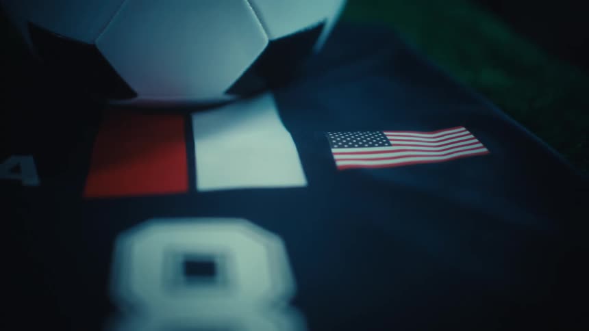A closeup shows a soccer jersey with a ball and small national flags including the united states softly lit against a dark background