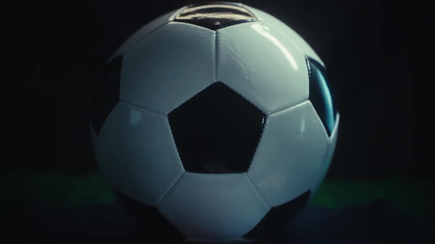 A close up shot of a classic black and white soccer ball resting on green artificial turf under dramatic dark lighting