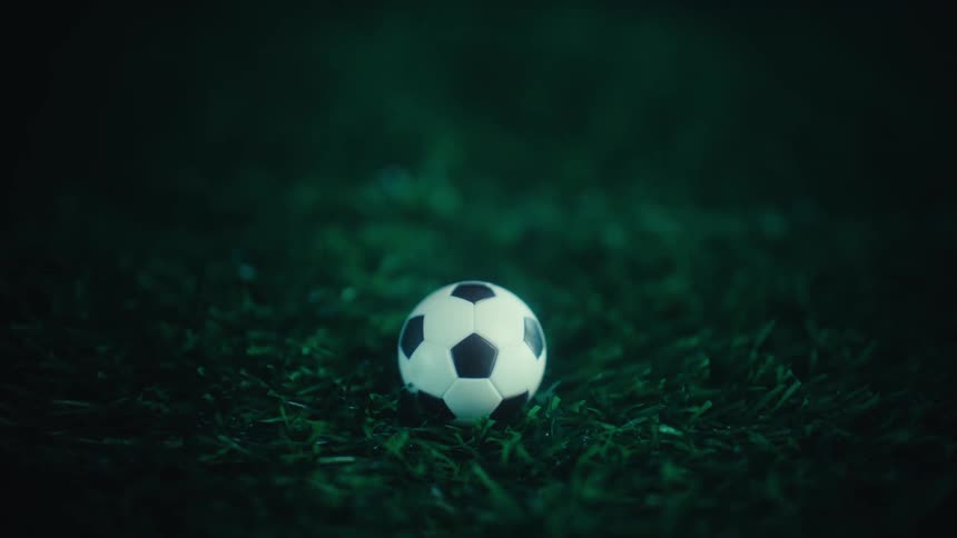 A soccer ball slowly comes into focus while sitting stationary on a dark green grass field under a moody cinematic spotlight at night emphasizing the texture of the turf and the classic black and white pattern of the ball
