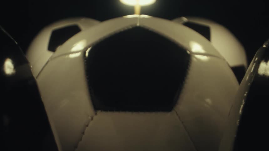 Several black and white soccer balls are clustered together under warm overhead lights in a dark studio setting