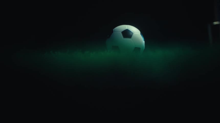 A soccer ball emerges from the darkness softly illuminated and partially obscured by a green haze