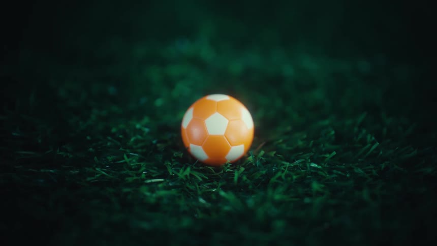 A close up shot of a small orange and white patterned soccer ball resting on green artificial turf under dramatic dark lighting