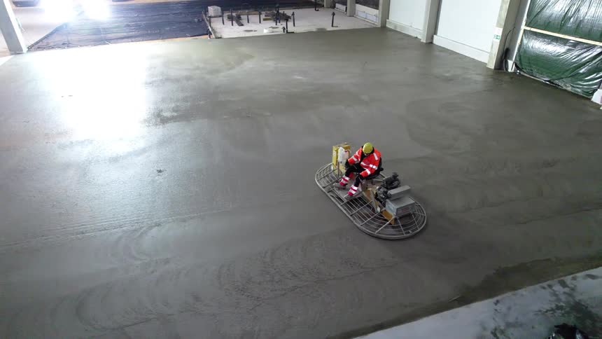 Worker with helmet operates trowel on concrete floor inside construction site