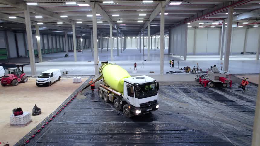 A large concrete mixer truck pours material inside a vast warehouse as workers prepare and oversee the construction site