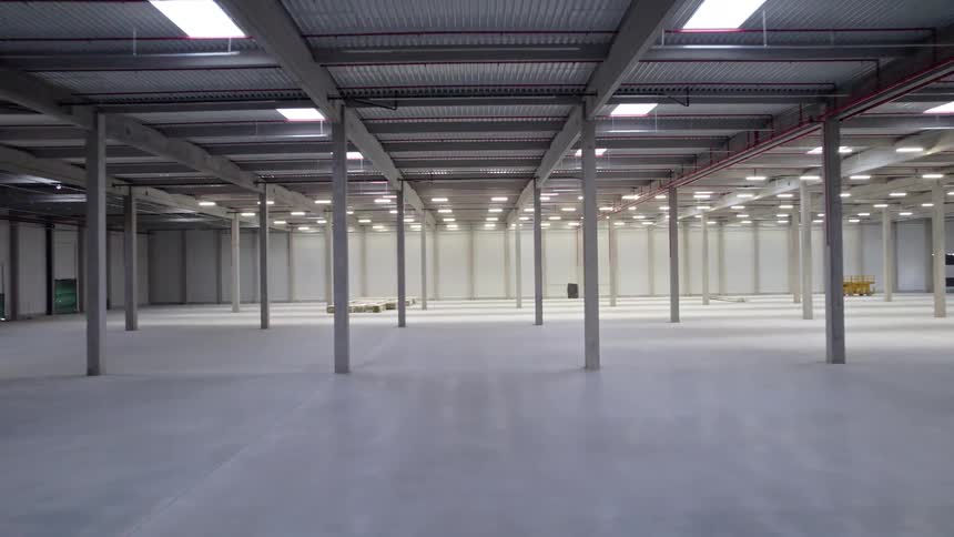 The view accelerates forward through a vast empty warehouse rushing past rows of concrete pillars under a ceiling of bright industrial lights