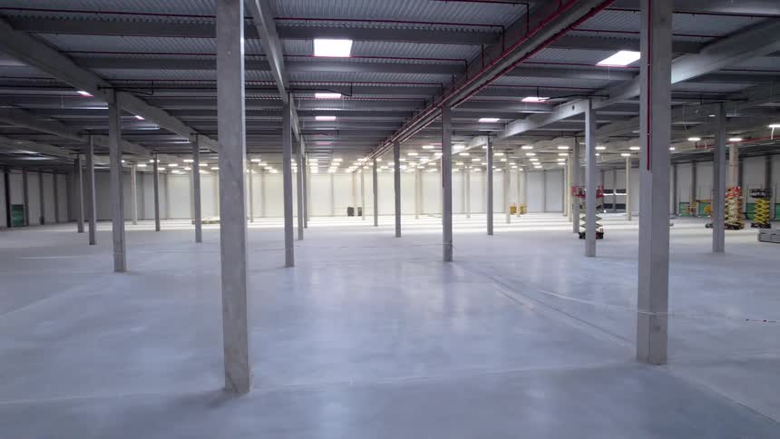 The camera slowly pans across the vast brightly lit interior of a massive empty warehouse showing rows of concrete pillars stretching into the distance