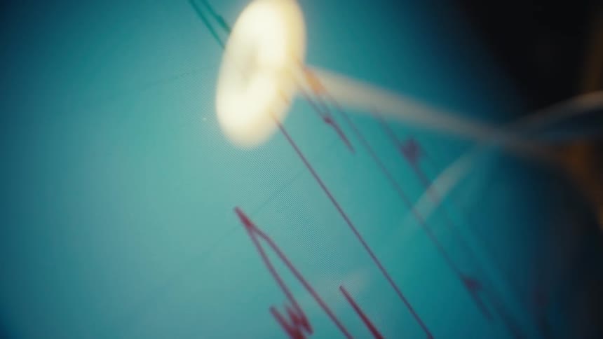 A closeup angled shot shows a glowing digital monitor displaying a green and red candlestick stock market chart as a desk lamp reflects on the screens surface in a dark room